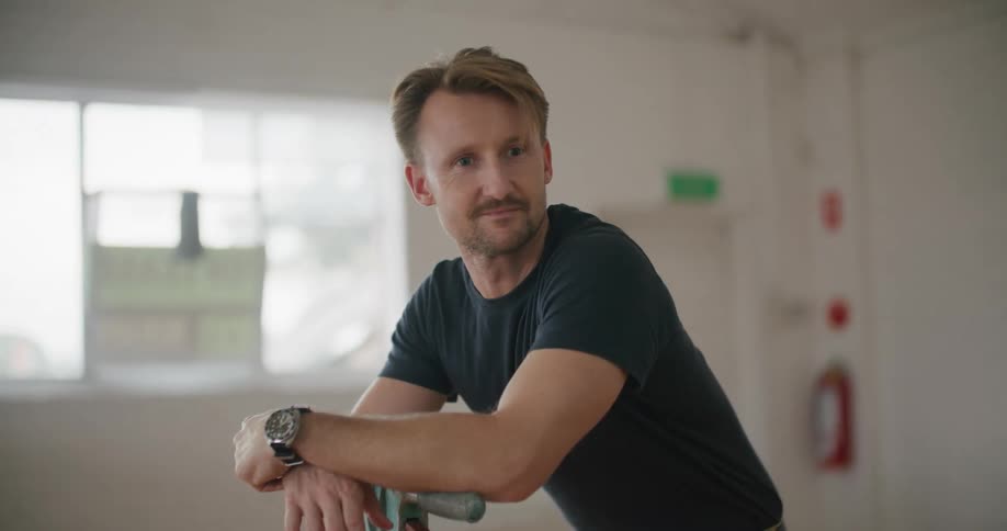 A man leans against a cleaning device in a room at daytime while looking around thoughtfully and smiles
