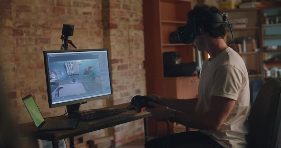 A man in a room sitting on a chair in front of a desk with a laptop and computer with a virtual reality headset on