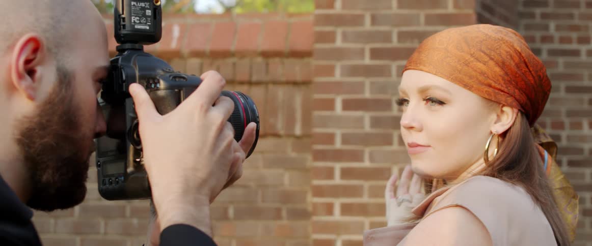 A man with a camera taking a picture of a woman with a wall trees the sky and clouds in the background