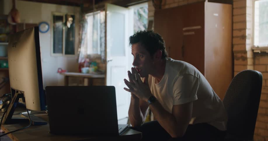 A man sits with his desk chair pushed out and his head in his hands as he looks at his laptop screen