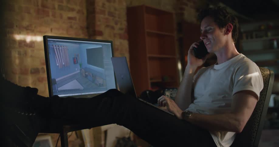 Man leaning back in chair at desk types on laptop while viewing larger monitor and talking on mobile phone
