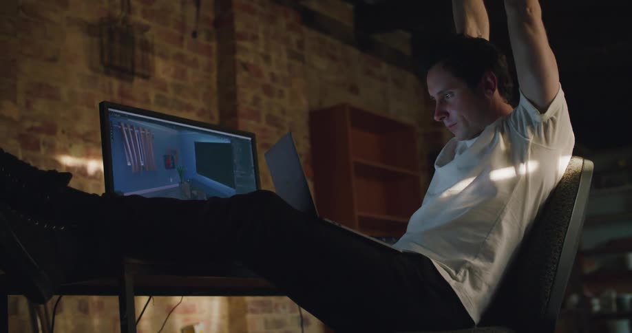 A man in a room sitting on a chair in front of a desk with a laptop and computer