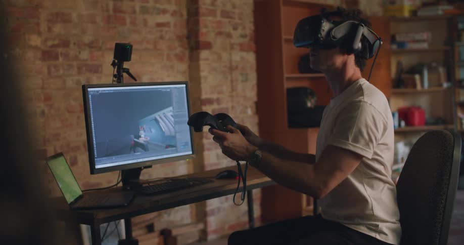 A man with a virtual reality headset sitting on a chair in front of a desk with a computer and laptop