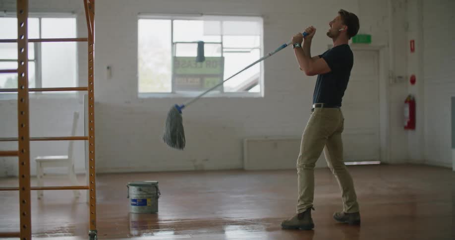 A man mopping in a room with a wooden floor dances around playfully and uses the mop as a mock mic during the day