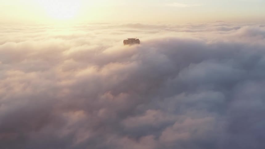 Aerial view of the sun rising over a blanket of clouds where a single building protrudes from the surface