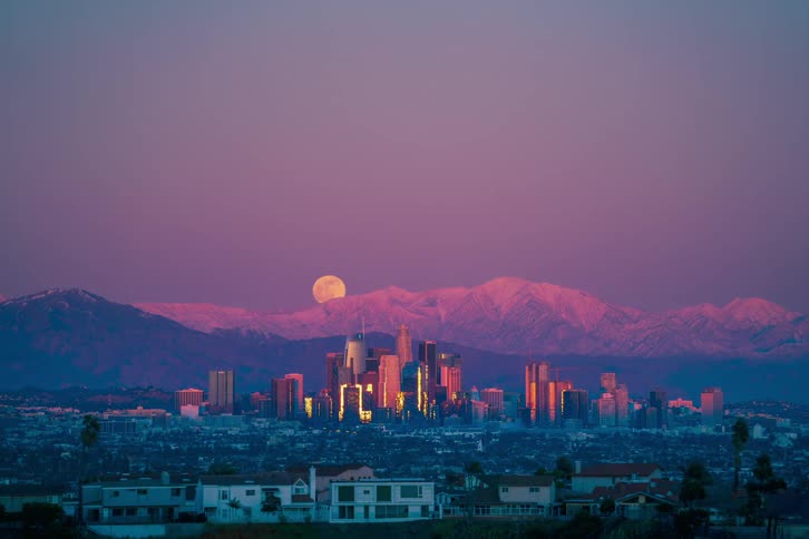 A city skyline with buildings with a mountain range the moon the sky and clouds in the background