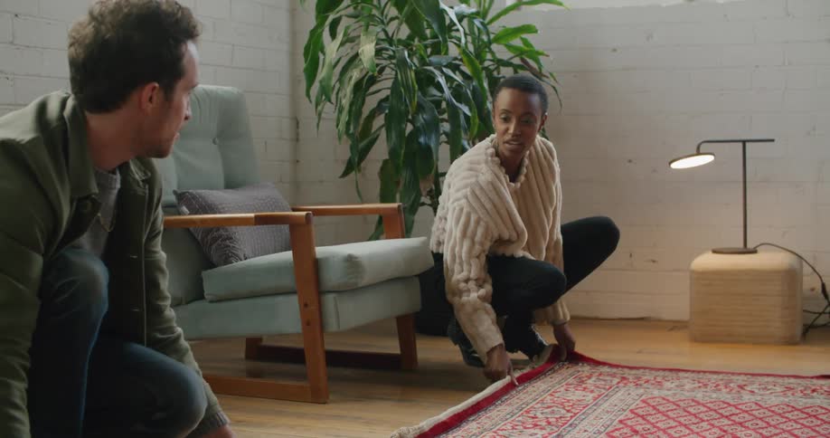 A man and woman on the floor in a room unrolling a carpet with a wall in the background