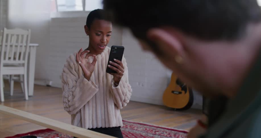 A woman in a room talking to a man while holding a smart phone with a guitar and a wall in the background