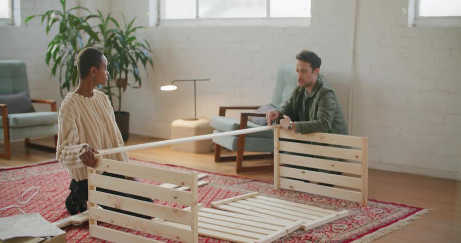 A woman and a man kneel down on a rug in a living room at daytime putting together a piece of wooden furniture