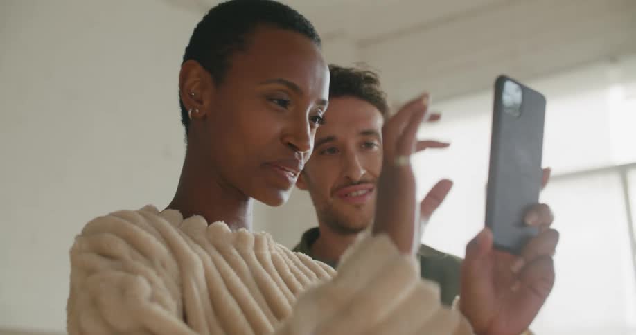 A man and a woman in a room looking at a smart phone with a wall and window in the background