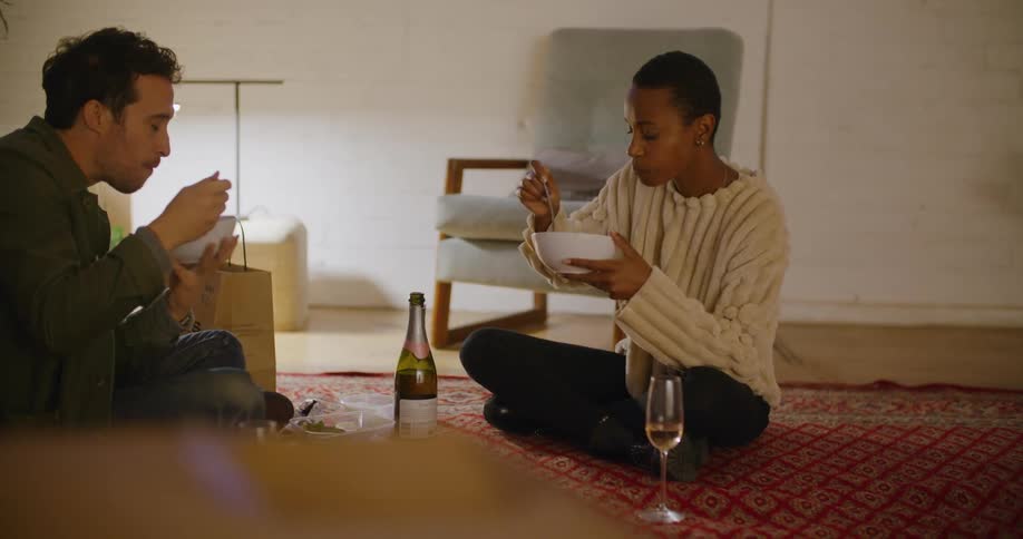 A man and a woman sitting on the floor eating with a wall and a plant in the background