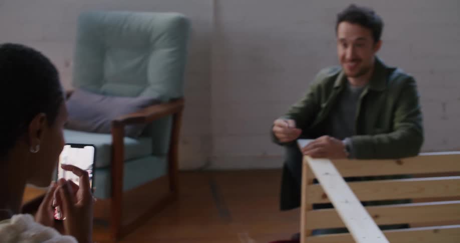 Woman watching instructions on her phone helps a man assembling furniture on the floor of the living room