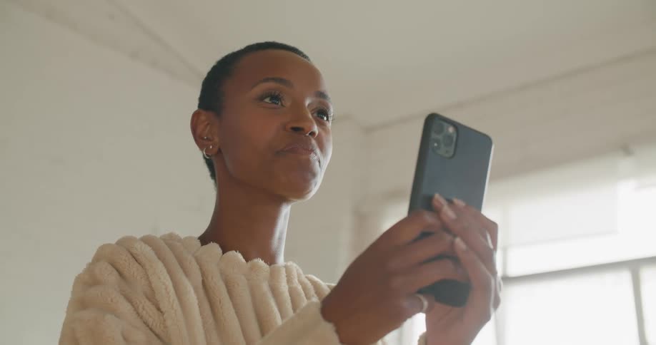 A woman in a room looking at a smart phone with a wall and window in the background
