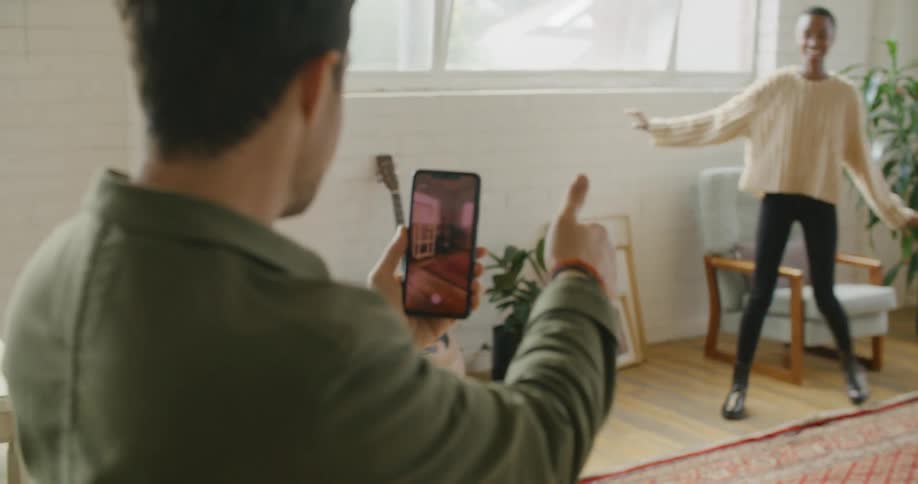 A man uses a smartphone to film a woman as she spins around on her feet in a house during the day