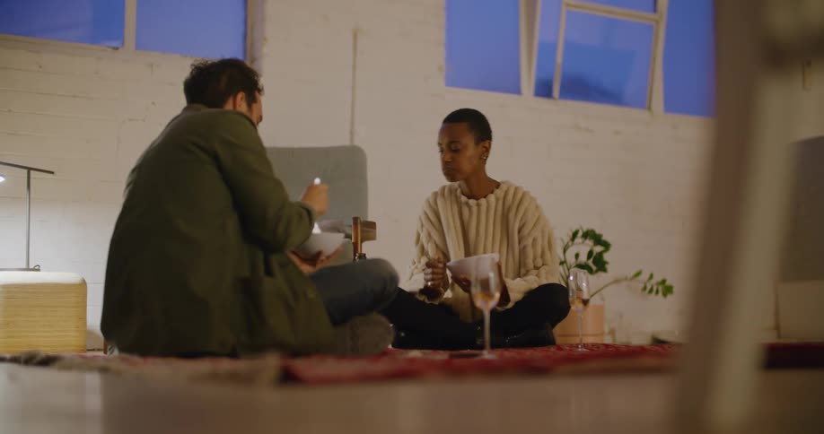 A man and a woman sitting on the floor eating with a wall and a plant in the background