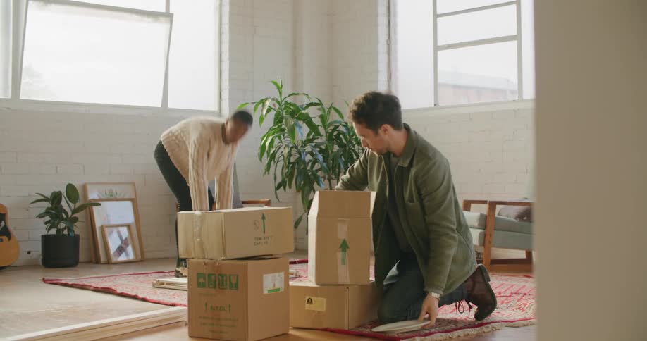 A man unpacks dishes from boxes as a woman moves wood across a room