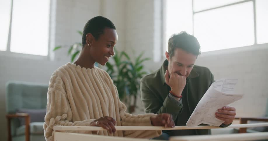 A man flips through an instruction manual as a woman next to him laughs while they kneel on a floor in a living room in front of an unfinished piece of furniture