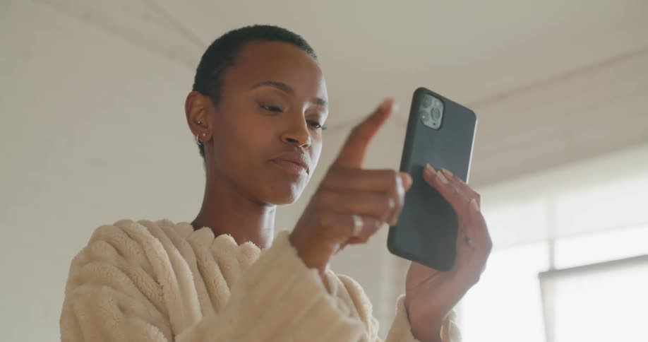 A woman in a room talking on a smart phone with a wall and window in the background