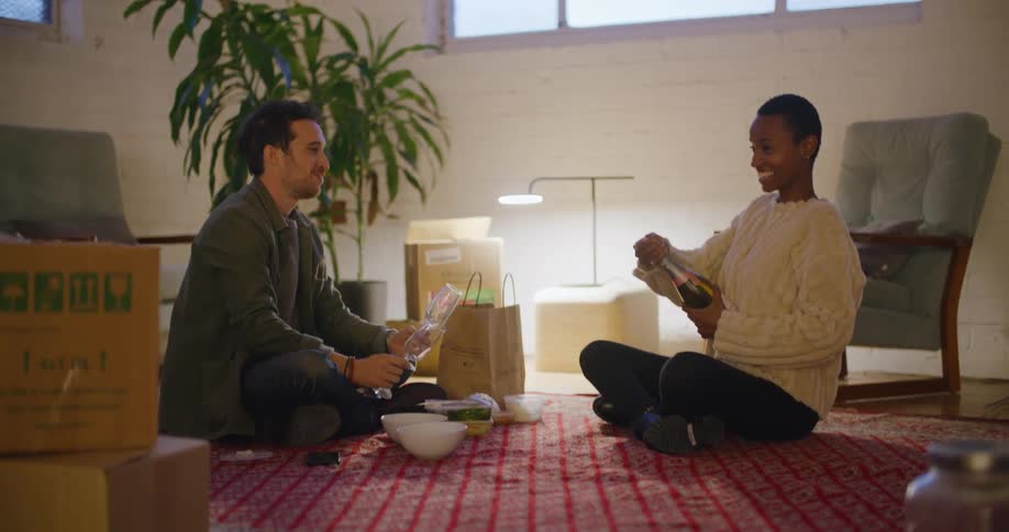 A woman opens a bottle of champagne and smiles with the man sitting on the floor holding flutes