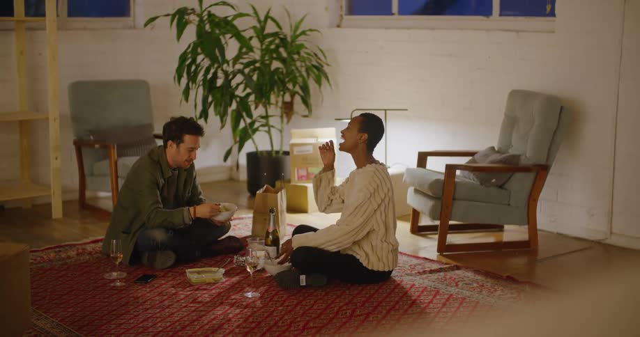 A young man and woman speak to one another as they eat while sitting on the floor of a house at night