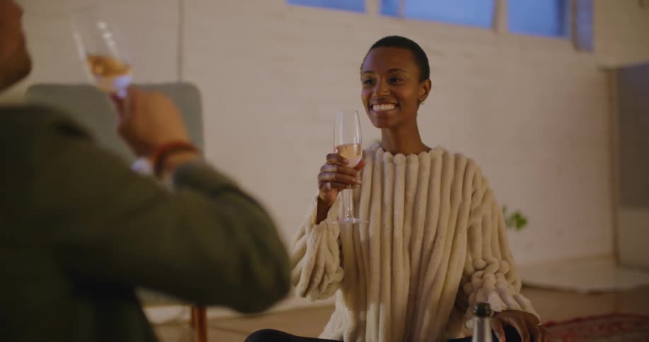 A woman sitting down on a living room floor pours wine into a wine glass and then makes a toast with a man sitting in front of her