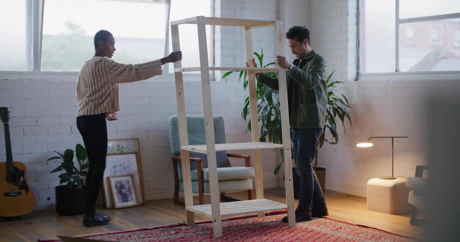 A man and woman lift a wooden shelf and move it as they decorate their apartment