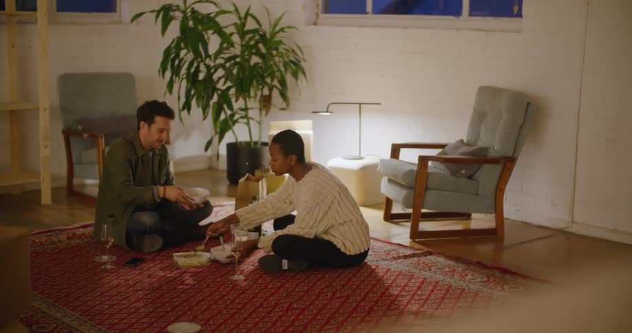 A couple sit on the floor of their living room and eat dinner out of white bowls