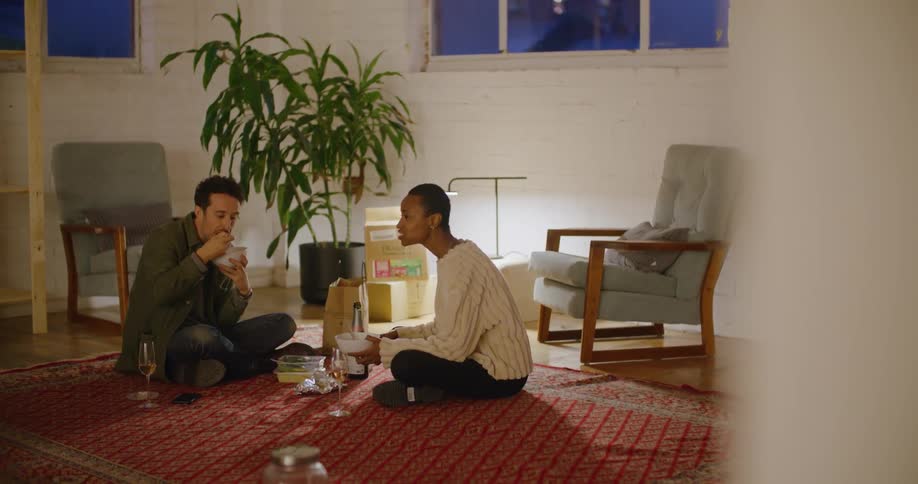 A man and a woman sitting on the floor eating with walls and windows in the background