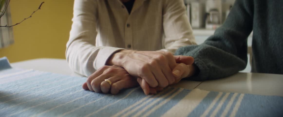Husband and wife stroke each others hands with thumb while sitting at table holding hands