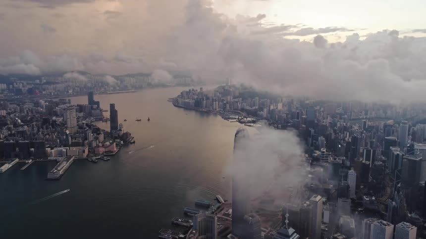 Low lying clouds flow across city skyscrapers bordering river as boats leave wakes