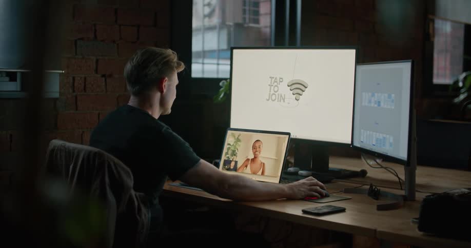 Man sitting at the desk inside a dimly lit office working on a computer while chatting on a video call