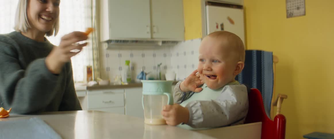 A woman sitting on a chair at a kitchen table feeding a baby with a wall and window in the background