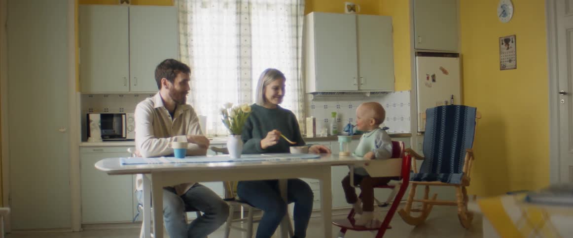 Family sits at kitchen table while mother feeds toddler and father drinks coffee
