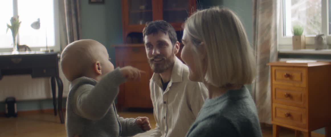 Mother turns head to look in direction toddler points as she and father sits on ground playing with child