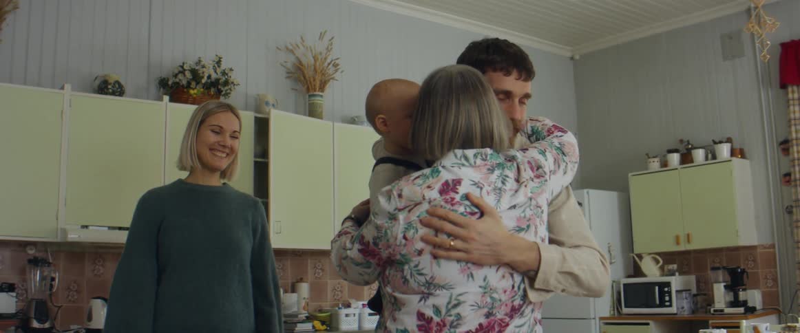 A husband and wife walking into a kitchen and greet an elderly woman as they hold a baby