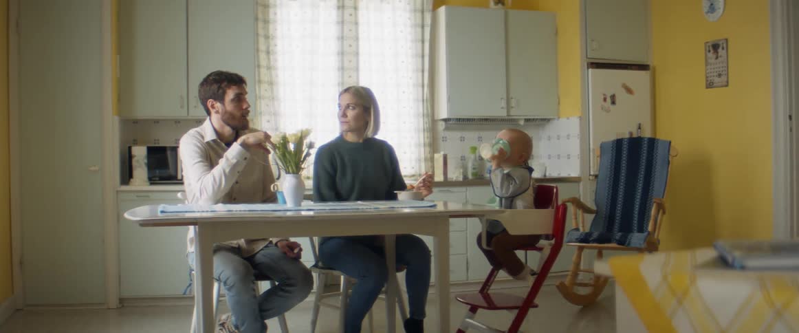 A couple sit at the kitchen table with their infant seated in a highchair while eating and drinking