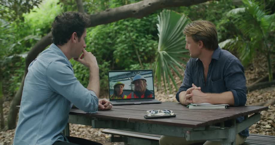 Two men sitting at a picnic bench and table looking at a laptop with trees and plants in the background