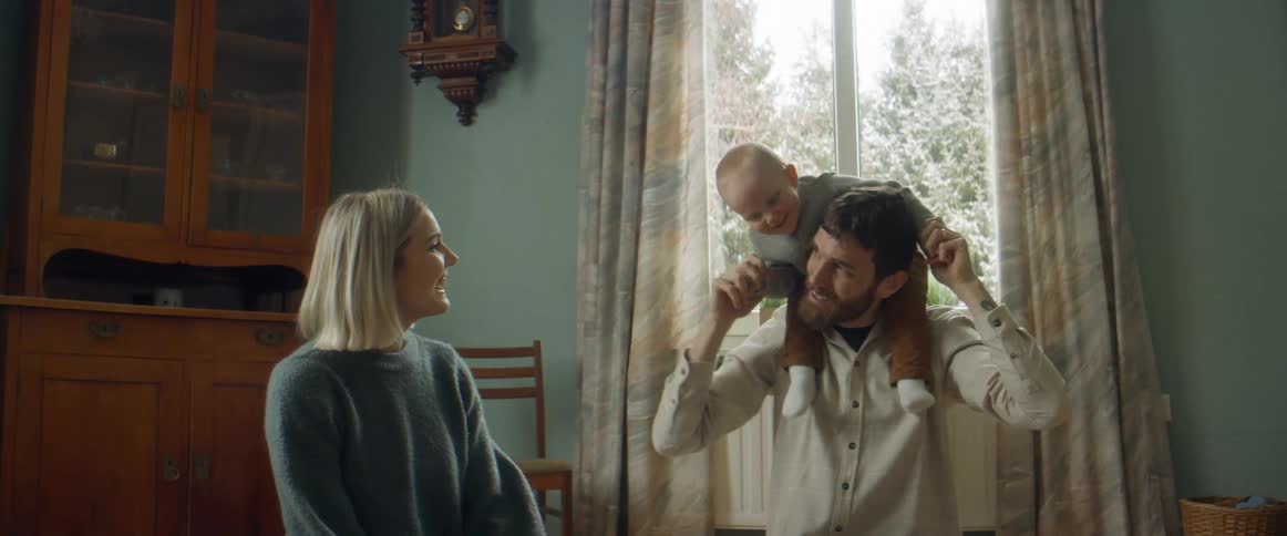 A husband and wife sit on the floor while the husband has their infant daughter seated on his shoulders