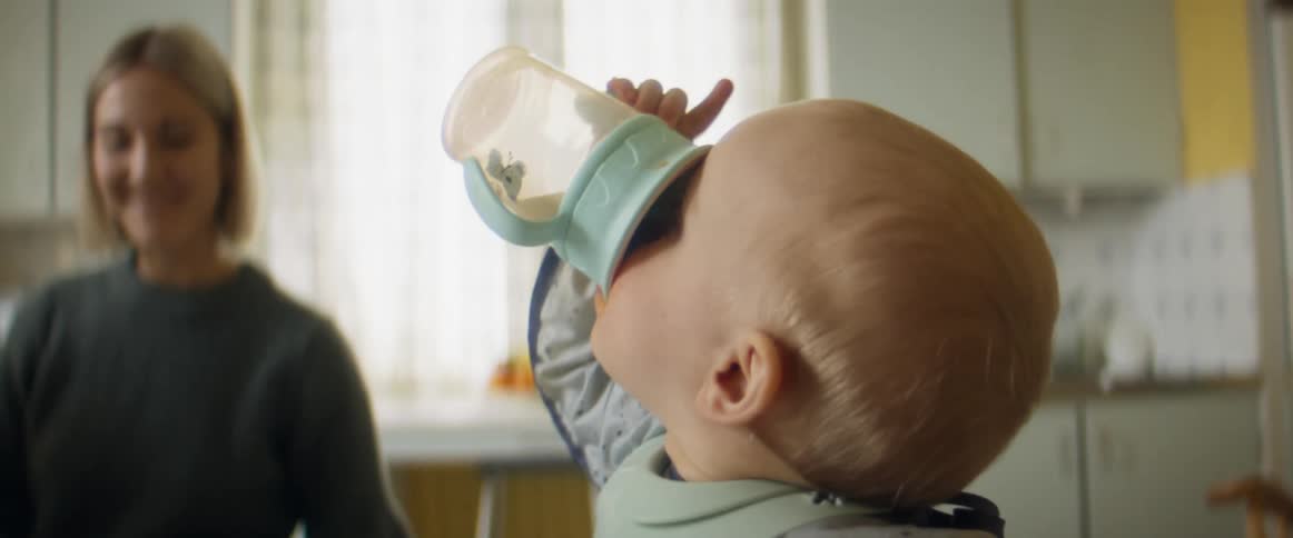 A young boy in a highchair grabs a sippy cup and takes a long drink as his mom sits down and smiles