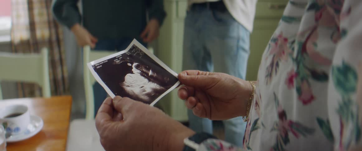 An older woman holds and looks at an ultrasound photo as a couple hold a baby nearby