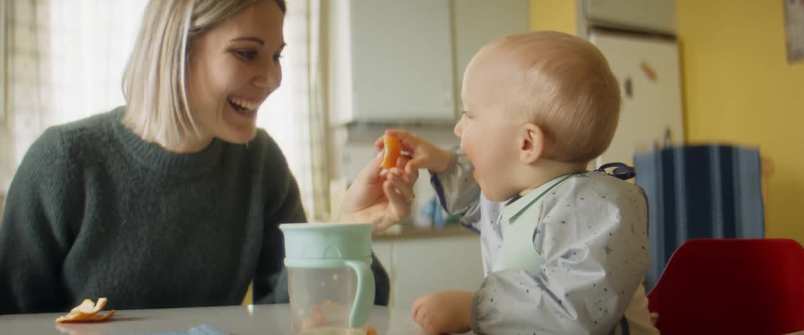 A woman smiles as she shows her infant son how to eat an orange as they sit around the kitchen table