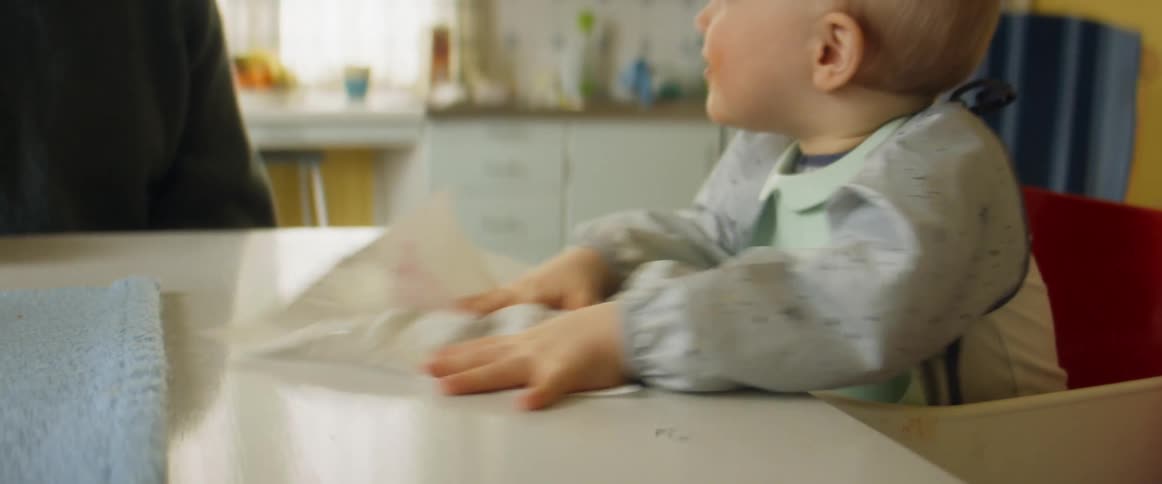 Mother watches toddler sitting in high chair wipe table