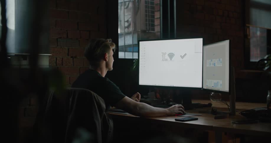 Man sitting in dark office manipulates graphics on large monitor at table set near brick wall and window