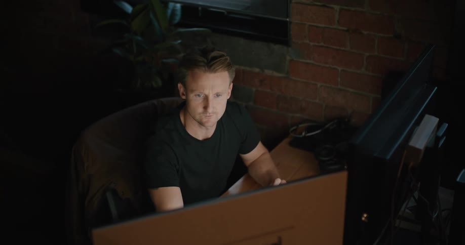 Man sitting at the desk inside a dimly lit room uses computers