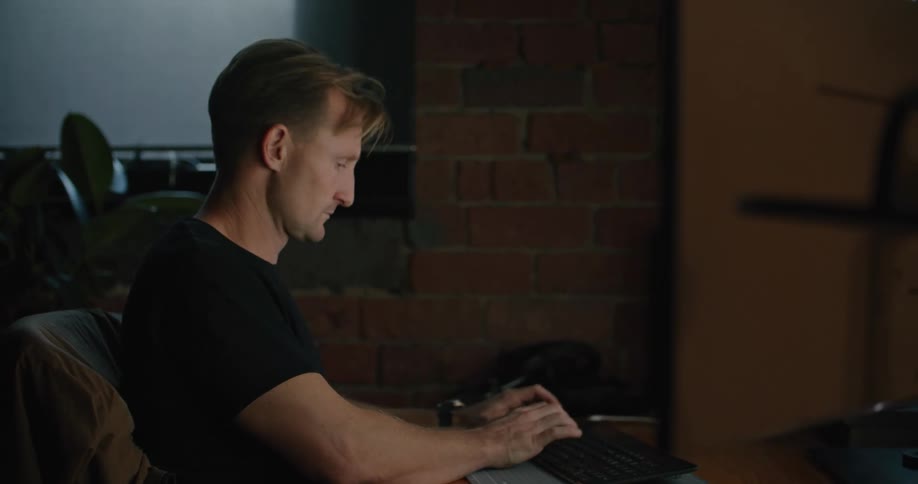 A man sitting on a chair at a desk typing on a keyboard with a wall and window in the background