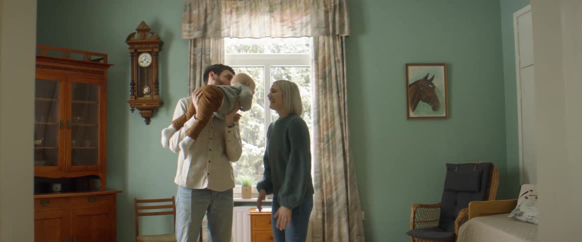 A man and a woman in a room holding and playing with a baby with a wall and window in the background