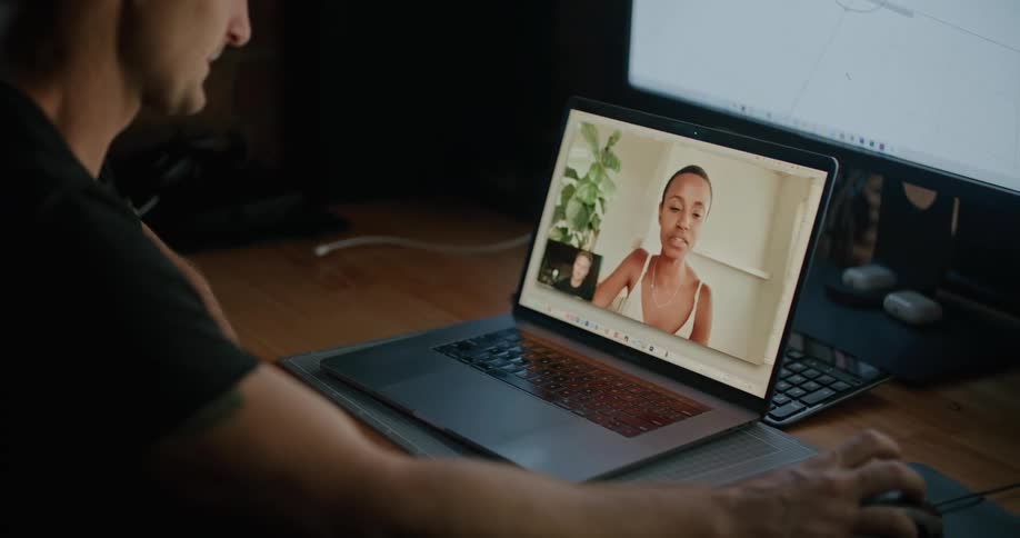A man sits at his wood desk and speaks to a colleague via video call on a laptop as another large monitor sits behind it