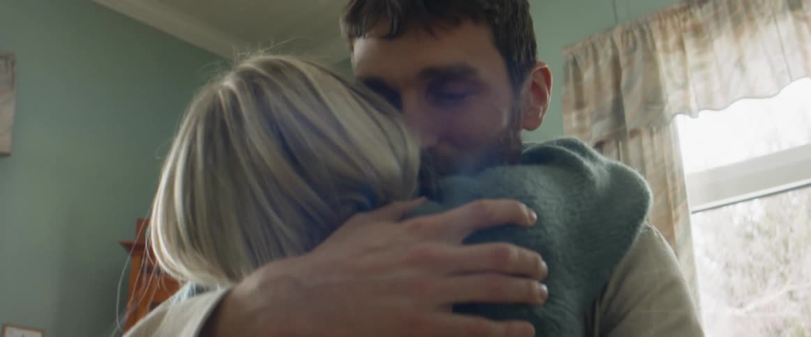 A man smiles and hugs his wife as they stand near a window in their house