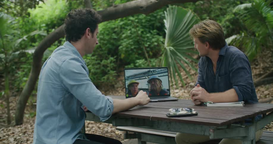 Men sit at picnic table having meeting via laptop with two onsite workers wearing hardhats