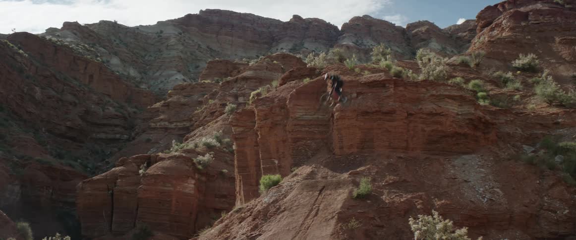A mountain biker spins their bicycle off a cliff before successfully landing on a dirt slope below